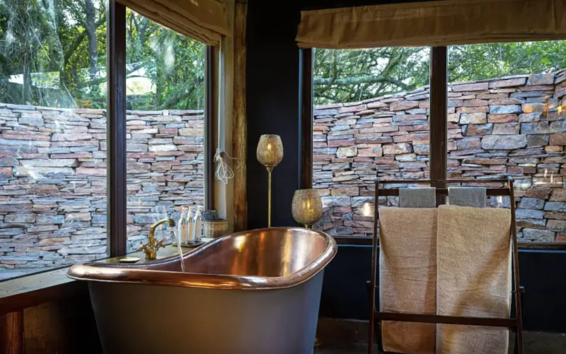 Luxurious copper bathtub in eco-luxury bathroom with stone walls, towels, lamps, and lush jungle views in Maasai Mara.