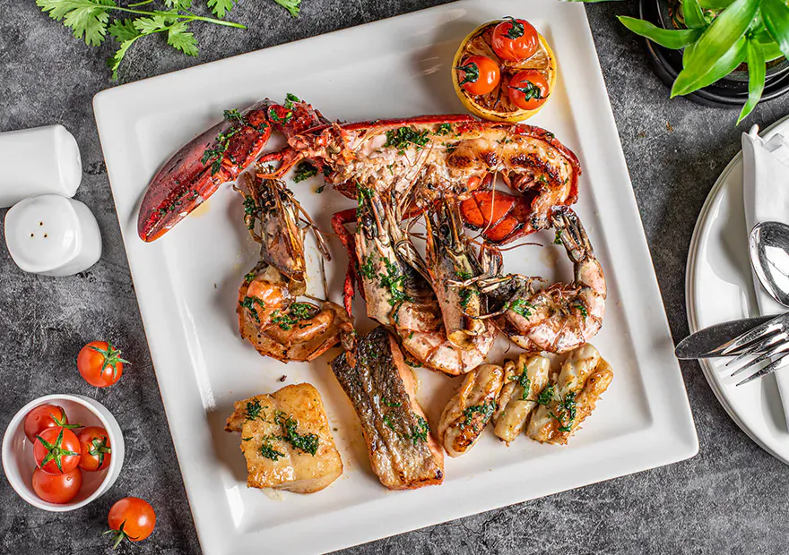 Square white plate of grilled lobster, prawns, fish, garnished with lemon slices, herbs on dark slate surface