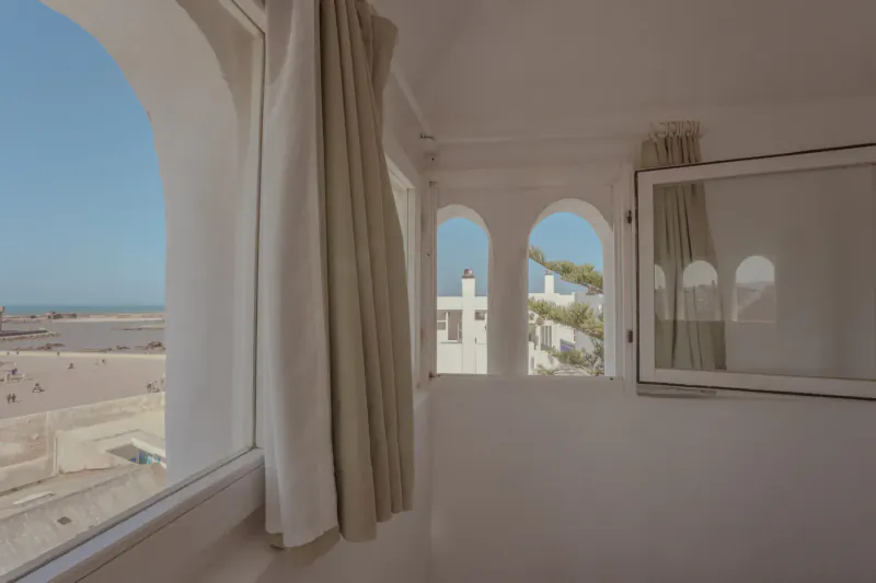 White arched windows with sheer curtains in Moroccan villa, framing sandy beach and ocean view in Essaouira.