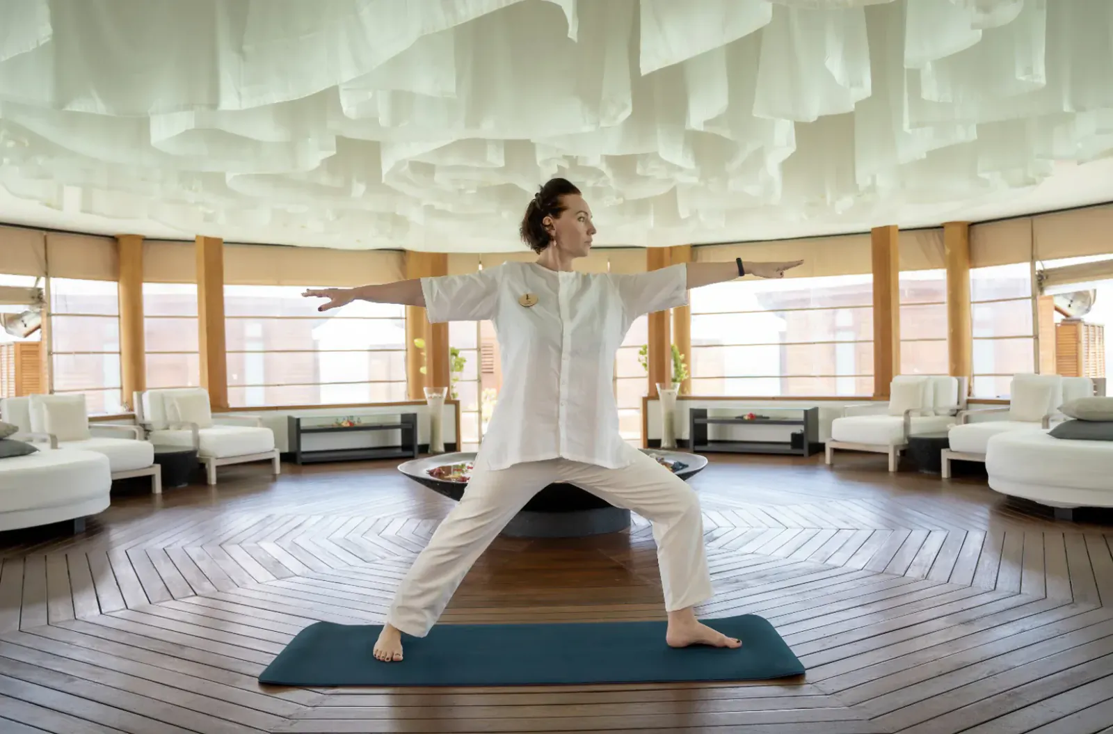 Woman in white outfit performs warrior yoga pose on mat in luxurious spa with wooden floors and lounge seating