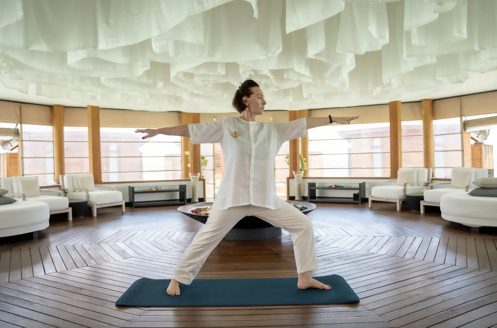 Woman in white outfit performs warrior yoga pose on mat in luxurious spa with wooden floors and lounge seating