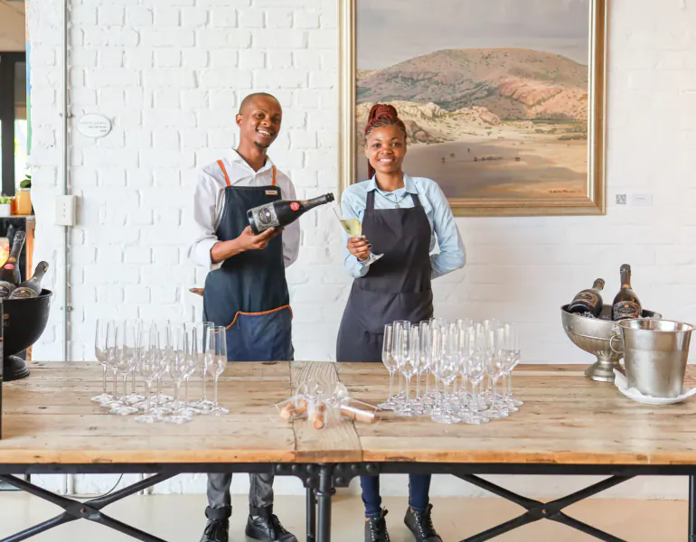Smiling Black man and woman in aprons hold champagne bottle and glass at rustic table with wine glasses and ice buckets in gallery.