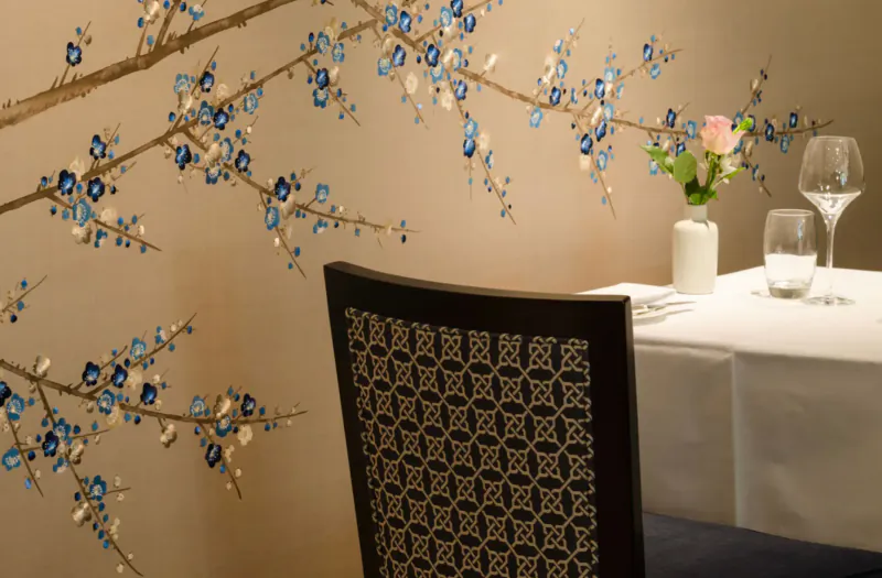 Elegant dining setup at Royal Crescent Hotel with blue blossom wall mural, patterned chair, white tablecloth, wine glasses, and pink rose.