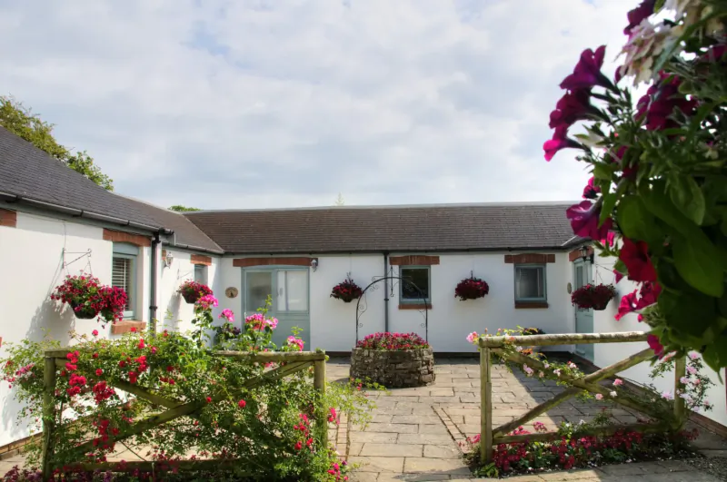 Idyllic courtyard at Broomhill Manor with white buildings, hanging flower baskets, central flower-filled well, and blooming gardens under partly cloudy sky.