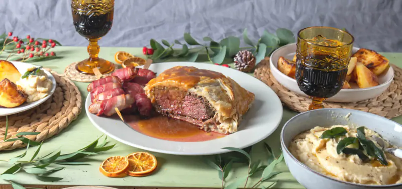 Christmas dinner box with beef Wellington, skewers, roast potatoes, dip, garnished with holly and oranges on green table.