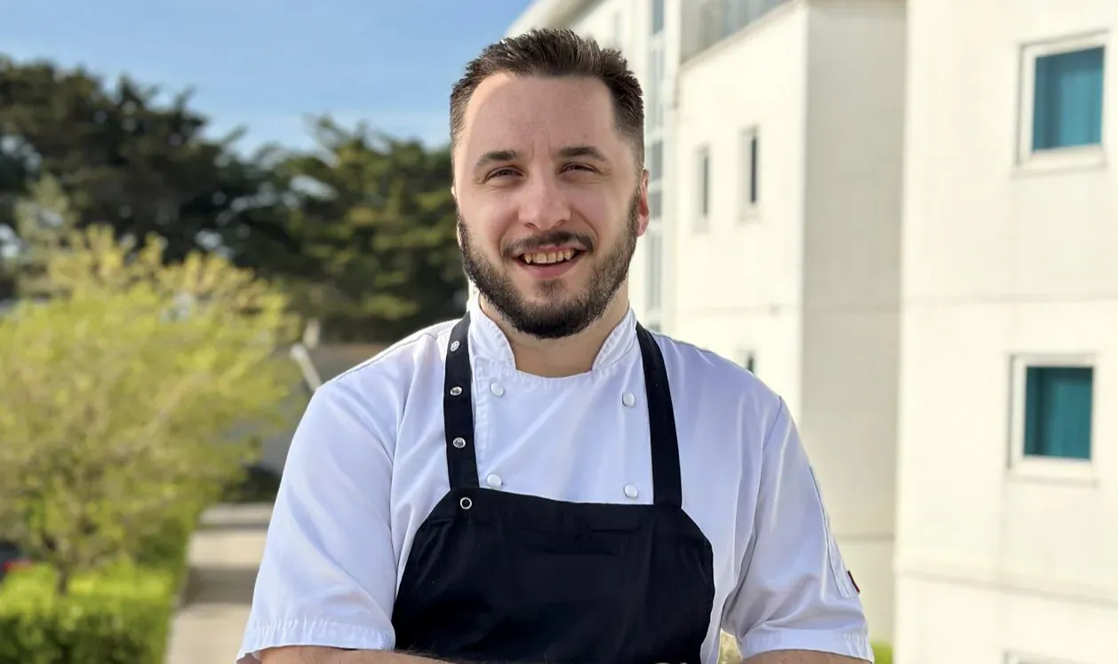 Chef Jake Grove smiling in white chef's jacket and black apron outside St Moritz Hotel with ocean view