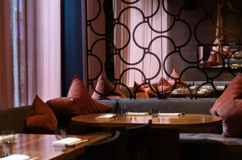 Elegant restaurant interior with round-patterned divider, orange pillows on gray sofa, and round wooden dining tables set with white napkins.