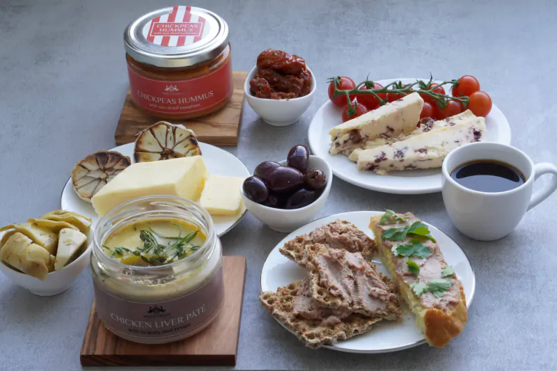 Picnic spread with Maison d'Entremets jar, cheeses, olives, tomatoes, artichokes, bread, and coffee on gray table.