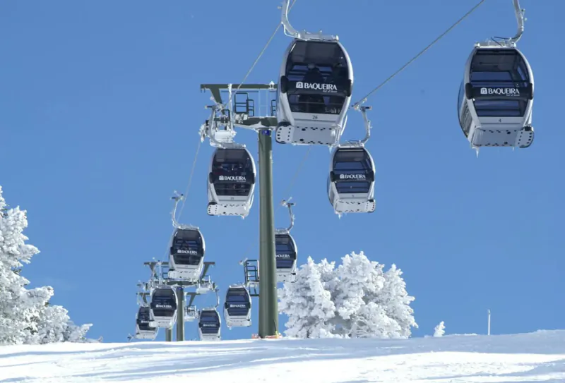 String of white gondolas branded 'Luxuria' ascending snowy ski slope amid frosted trees under blue sky