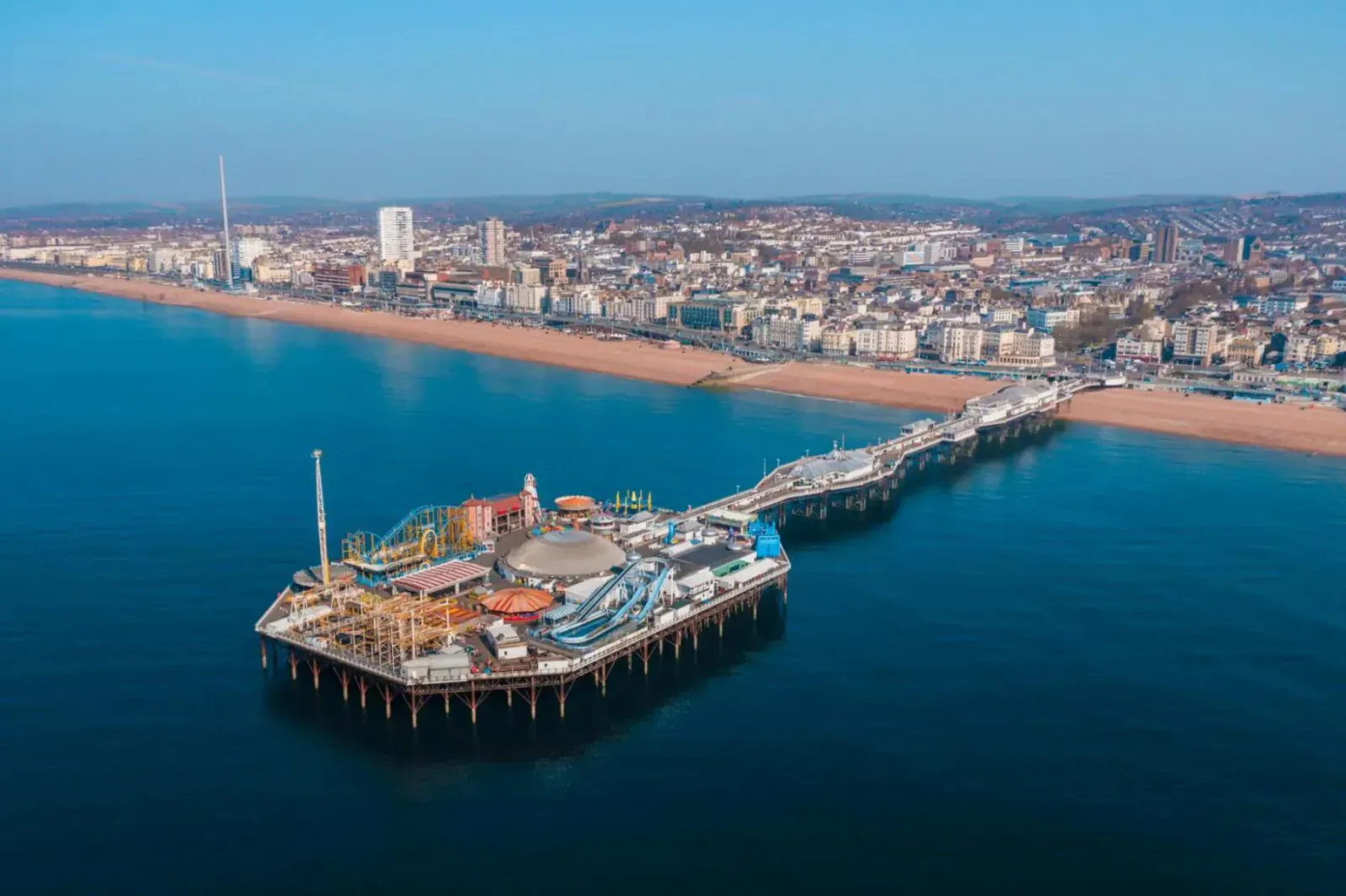 Aerial view of Brighton Palace Pier with rides extending over the sea and sandy beach, city skyline beyond.