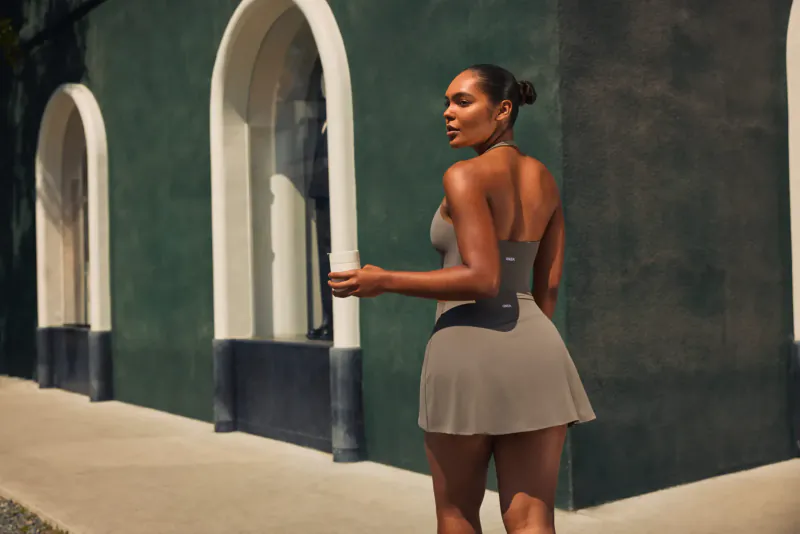 Black woman in gray backless crop top and mini skirt holds drink, poses by arched green building