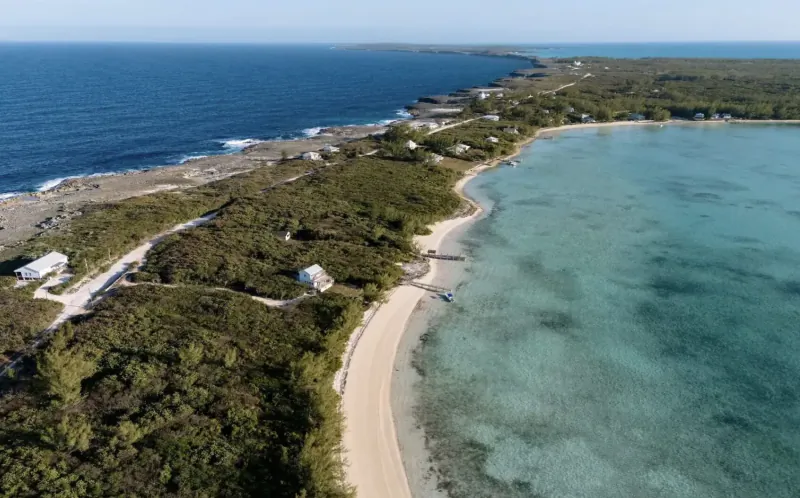Aerial view of Lot 43 Bottom Harbour Beachfront: sandy beach curving between turquoise lagoon and ocean, surrounded by lush greenery and white structures, Bahamas.