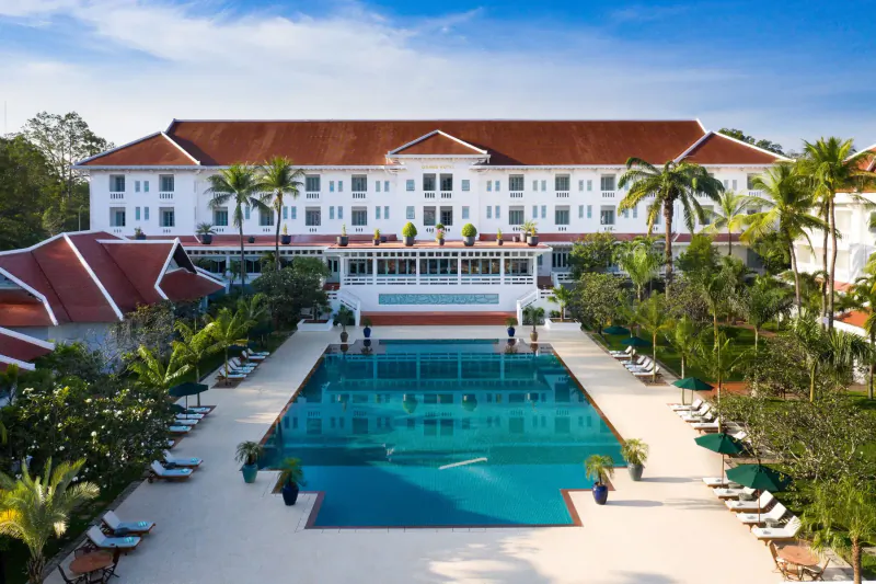 Aerial view of luxurious white colonial-style resort with red roofs, central turquoise pool, and palm trees.
