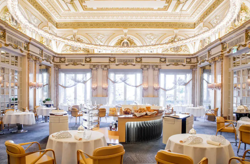 Opulent Michelin restaurant interior in Monaco with ornate gilded ceiling, large windows, and elegantly set round tables.