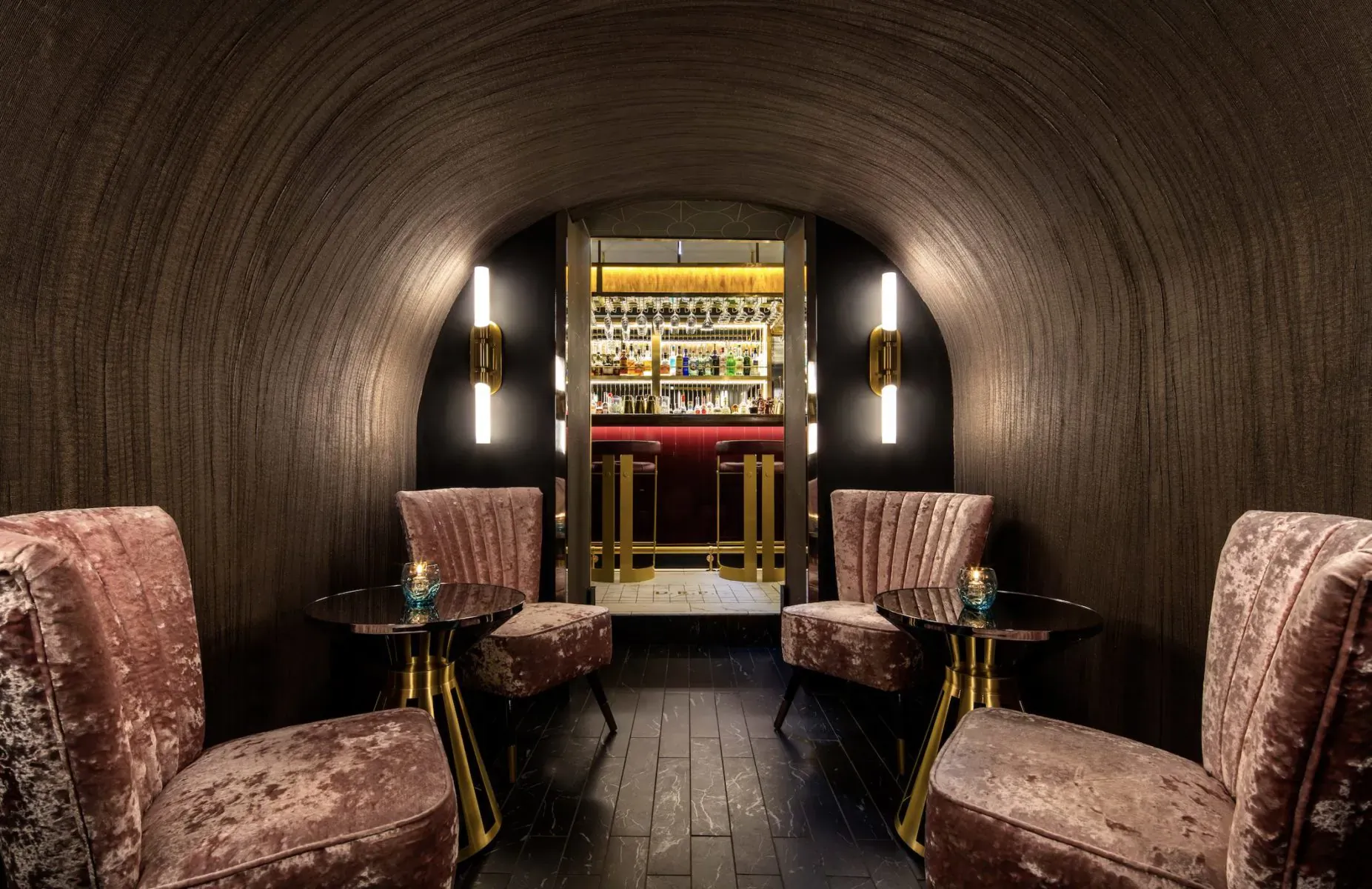 Cozy curved tunnel lounge at Disrepute Bar with plush pink velvet chairs, gold-accented bar stocked with bottles, and candles.