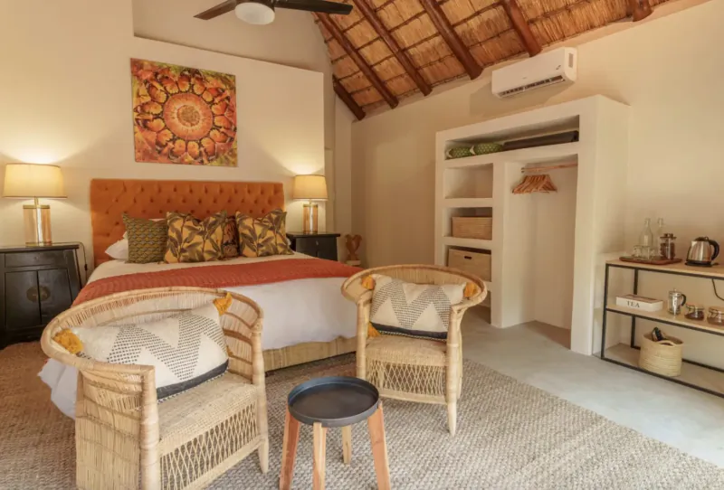 Cozy thatched-roof bedroom in wilderness lodge with orange bed, rattan chairs, vibrant mandala art, and tea station.