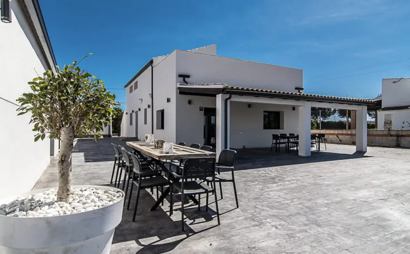 White modern villa with covered terrace, long wooden dining table with chairs, potted olive tree, on sunny pebbled patio
