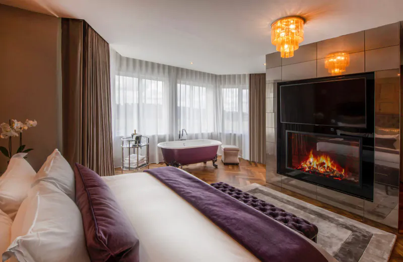 Luxurious hotel bedroom with king bed, pink bathtub, burning fireplace, and city view windows