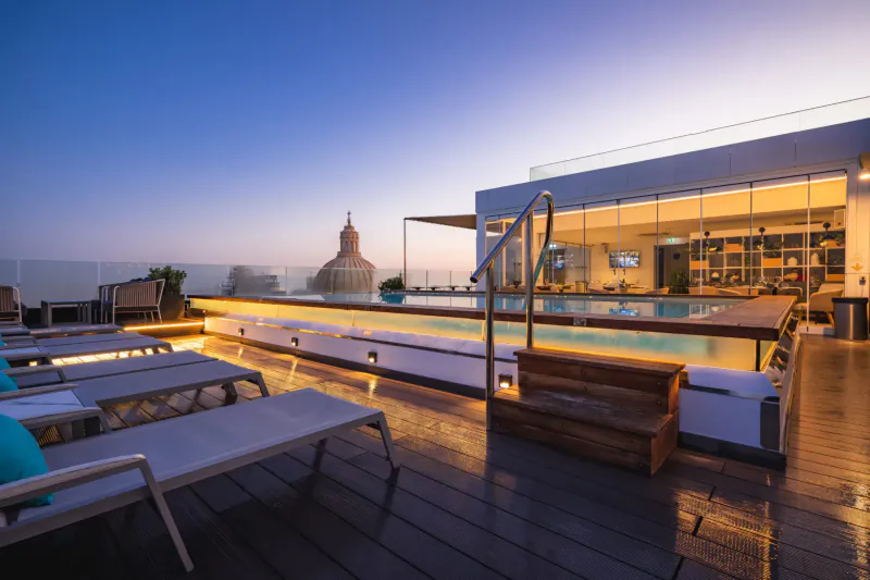 Twilight rooftop pool at Embassy Valletta Hotel with lounge chairs, modern bar, and distant golden dome in Malta