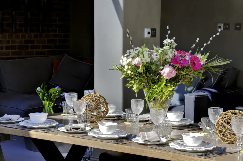 Elegant dining table in luxury cottage with white plates, glasses, colorful flower bouquet, rattan orbs, and lounge seating nearby