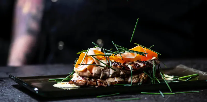 Stacked salmon with sunny-side-up eggs and microgreens on black plate, chef's tattooed arm in background