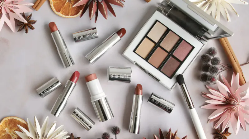 Assortment of MERIT silver lipsticks, eyeshadow palette, brush amid pink flowers, orange slices, star anise on white surface