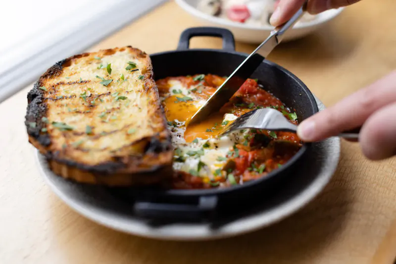 Hand slicing poached egg in shakshuka served with garlic bread in black pan on wooden table