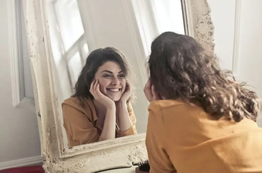 Smiling woman with curly hair in yellow top, hands on cheeks, reflected in white ornate mirror
