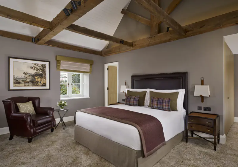 Elegant Langley Hotel bedroom with exposed beam ceiling, king bed with plaid pillows, leather armchair, and window.