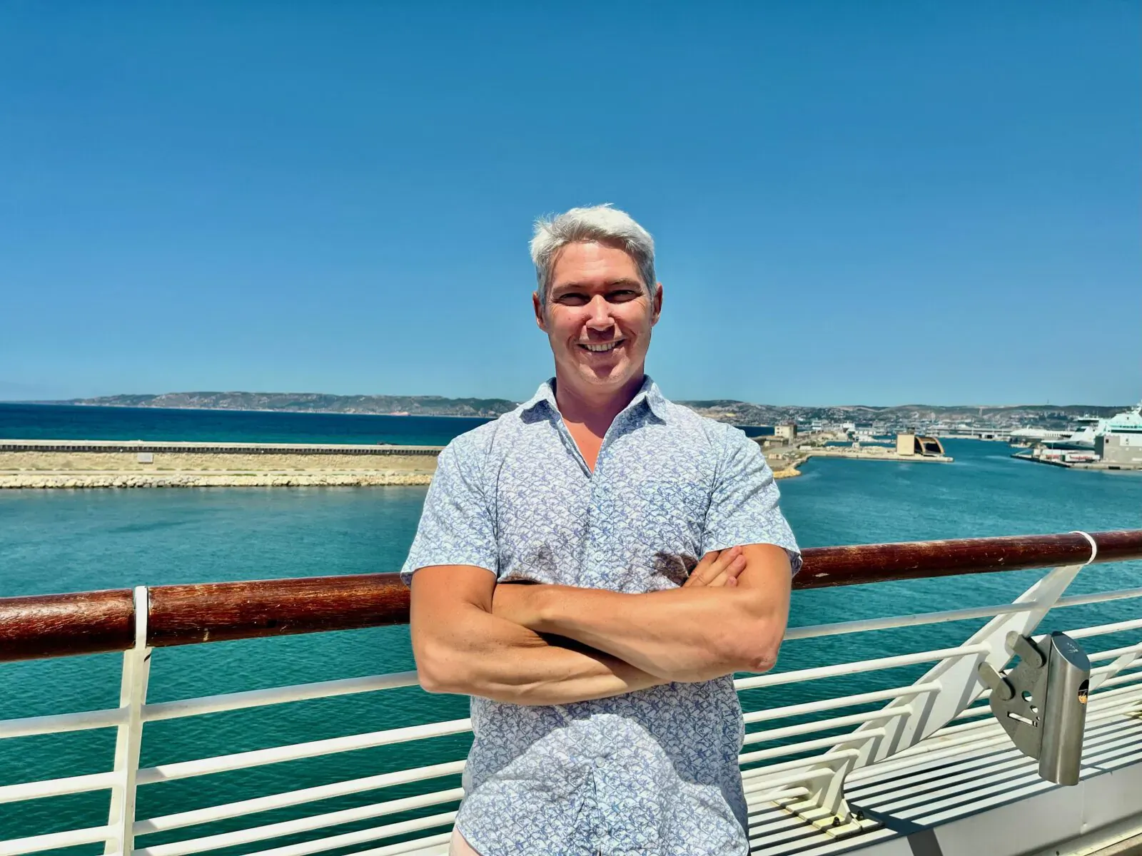 Mr. Theo Dandine, owner of Amber Lombok Beach Resort, smiles with arms crossed on cruise ship deck overlooking blue sea and port.