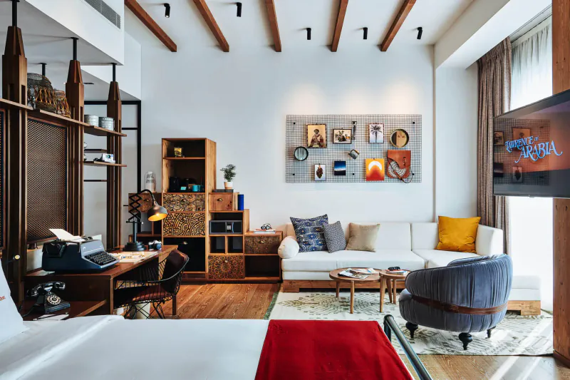 Spacious modern hotel room at 25hours Hotel One Central Dubai with wooden beams, art wall, desk, sofa, TV, and bed.