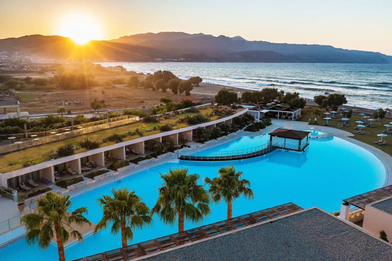 Aerial view of Cavo Spada Resort's curved blue infinity pool, palm trees, sun loungers at sunset with sea and mountains, Crete.