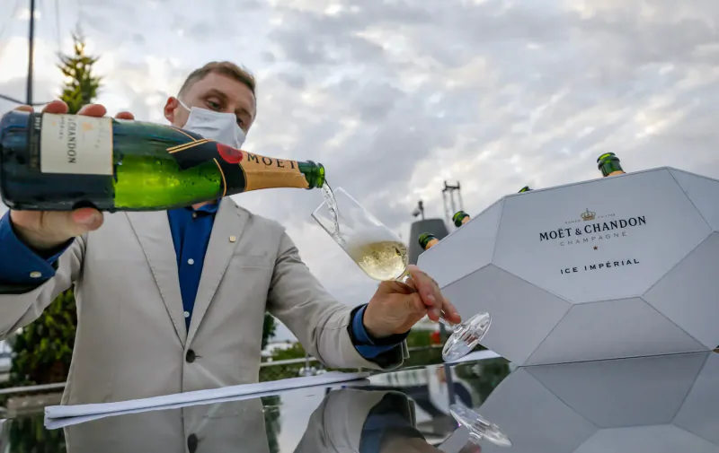 Man in suit and mask pours Moët & Chandon Impérial champagne from green bottle into flute at luxury event, with boxes nearby.