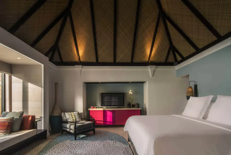 Luxurious Maldives villa bedroom with thatched ceiling, king bed, pink cabinet, TV, and chairs