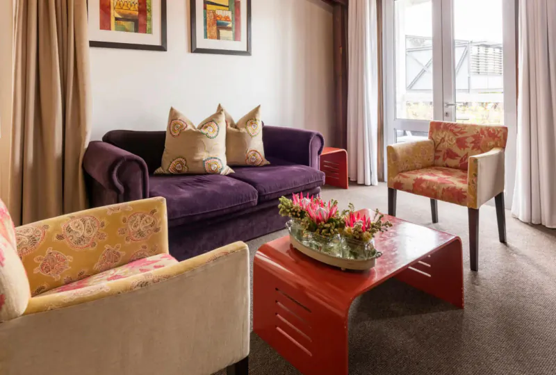 Cozy hotel living room with purple velvet sofa, floral armchairs, red coffee table with flowers, balcony view.