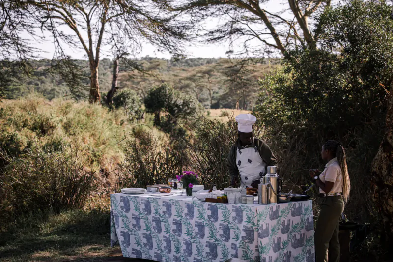 Chef in white uniform and toque serves food at outdoor table with woman, acacia savanna backdrop, El Karama Lodge.