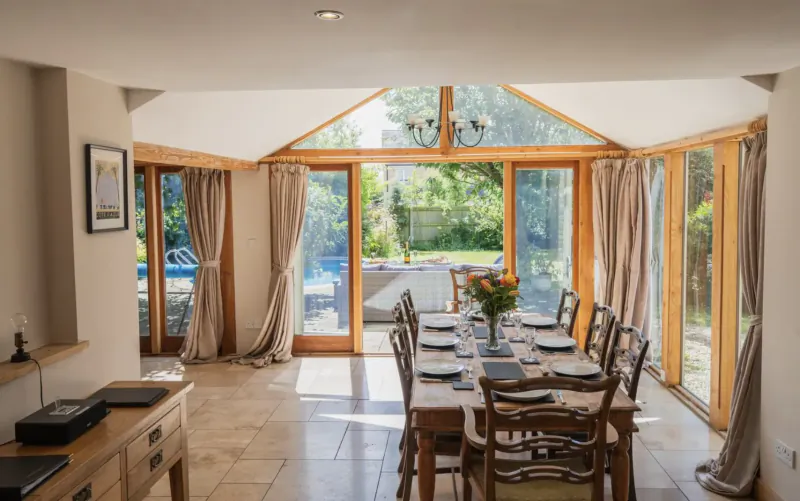Bright dining room in Cotswolds country house with wooden table set for meal, French doors to garden pool.