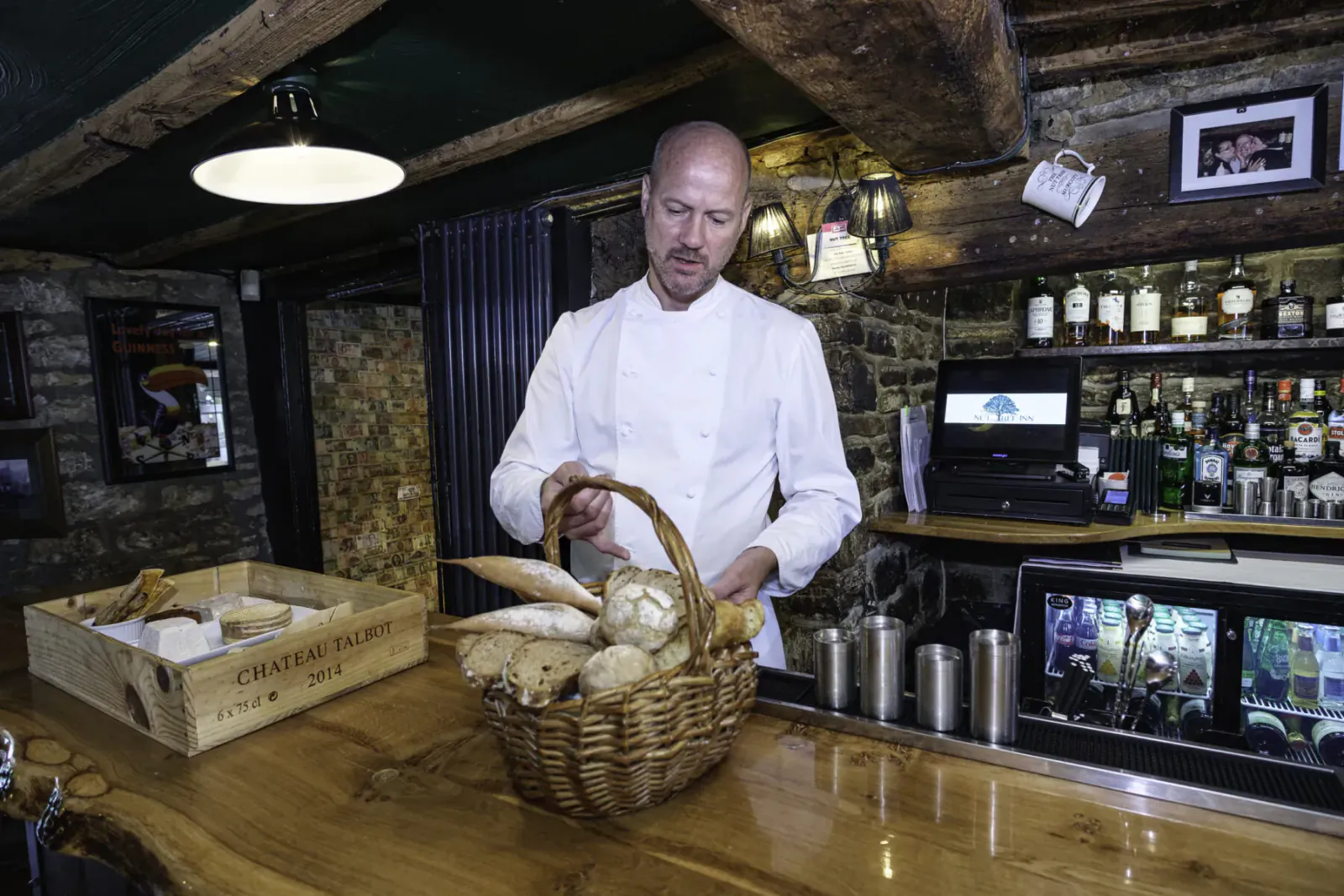 Bald chef in white uniform holding basket of baguettes over wooden bar at Nut Tree Inn, rustic stone interior with liquor bottles.