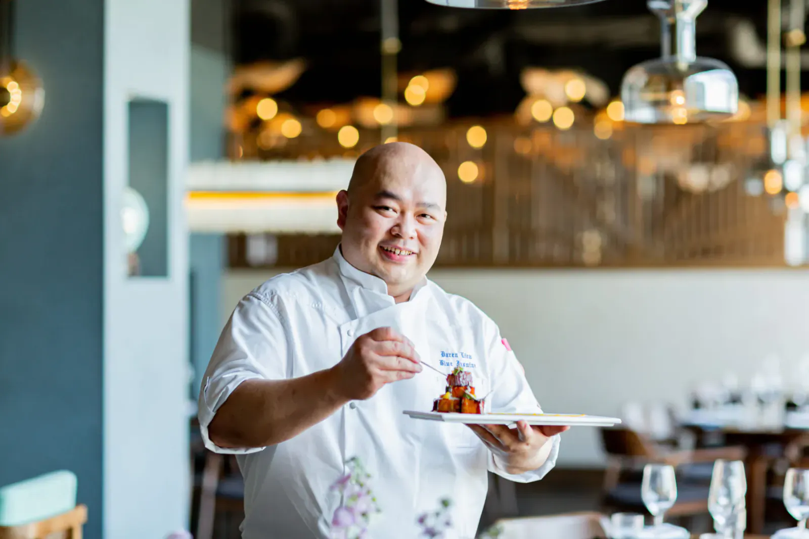 Bald smiling chef in white uniform holding plate of stacked dish in modern restaurant with pendant lights