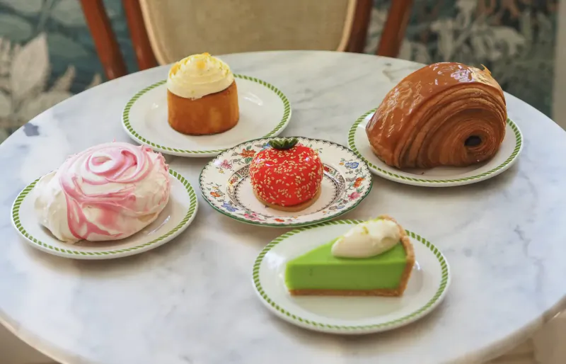 Assortment of pastries on plates on a marble table: pink meringue, red strawberry cake, green lime tart, yellow cupcake, brown croissant.