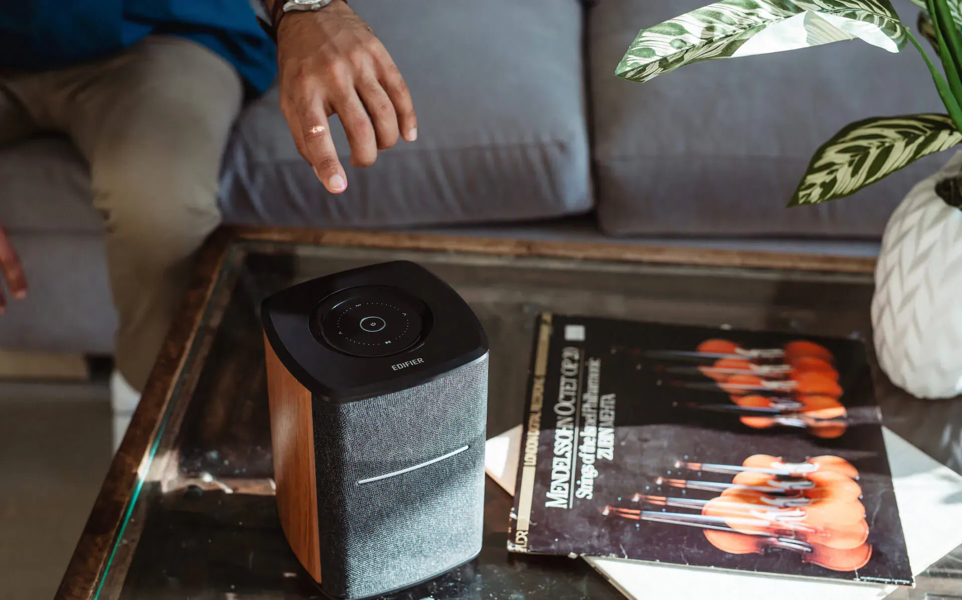Man's hand reaches toward new Edifier MS50A smart speaker on glass table with gray sofa, plant, and album nearby.