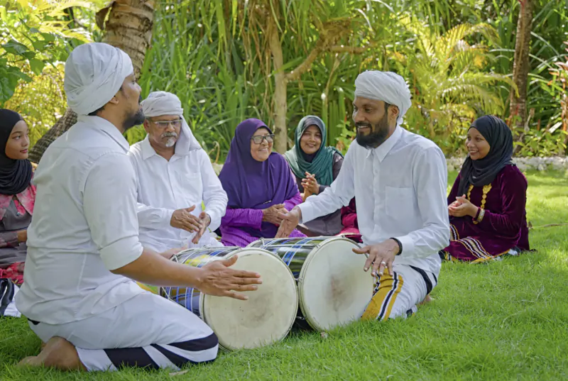 Maldivian men in white turbans and kurtas playing large drums, with women in hijabs, sitting on lush green lawn amid palms.