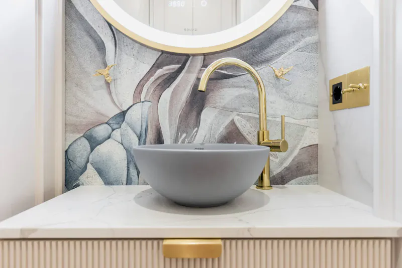 Modern gray vessel sink with gold faucet on white marble vanity, artistic floral wallpaper backdrop in luxury bathroom.