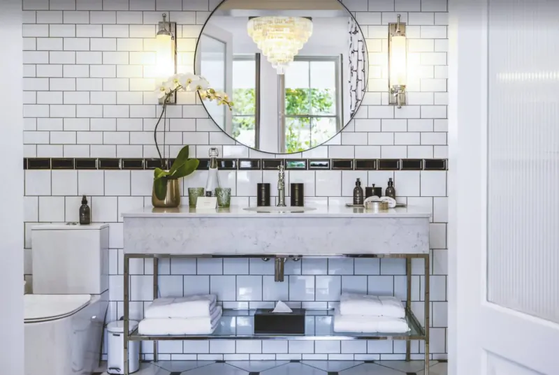 Elegant Cape Cadogan hotel bathroom with white subway tiles, marble vanity, orchids, chandelier, and round mirror.