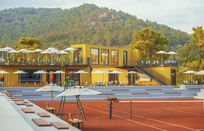 Yellow shipping container resort with terrace overlooking red clay tennis court and green mountains at Corendon Playa Kemer, Turkey