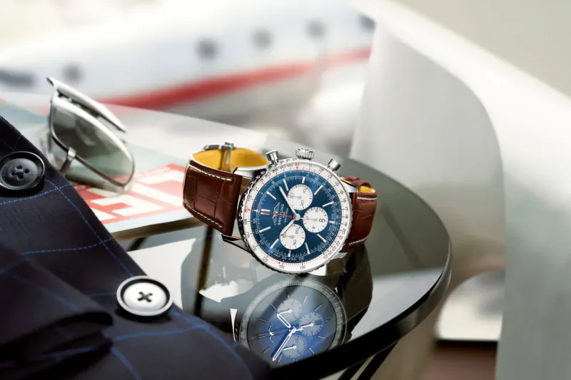 Breitling Navitimer blue chronograph watch with brown leather strap on glass table near airplane and jacket