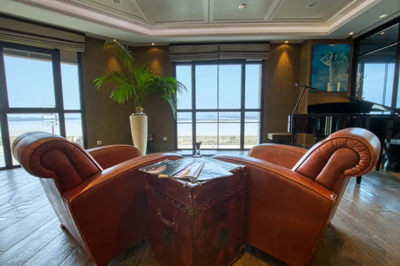 Luxurious hotel lounge with two leather armchairs, coffee table with magazines, grand piano, palm plant, and sea view windows.