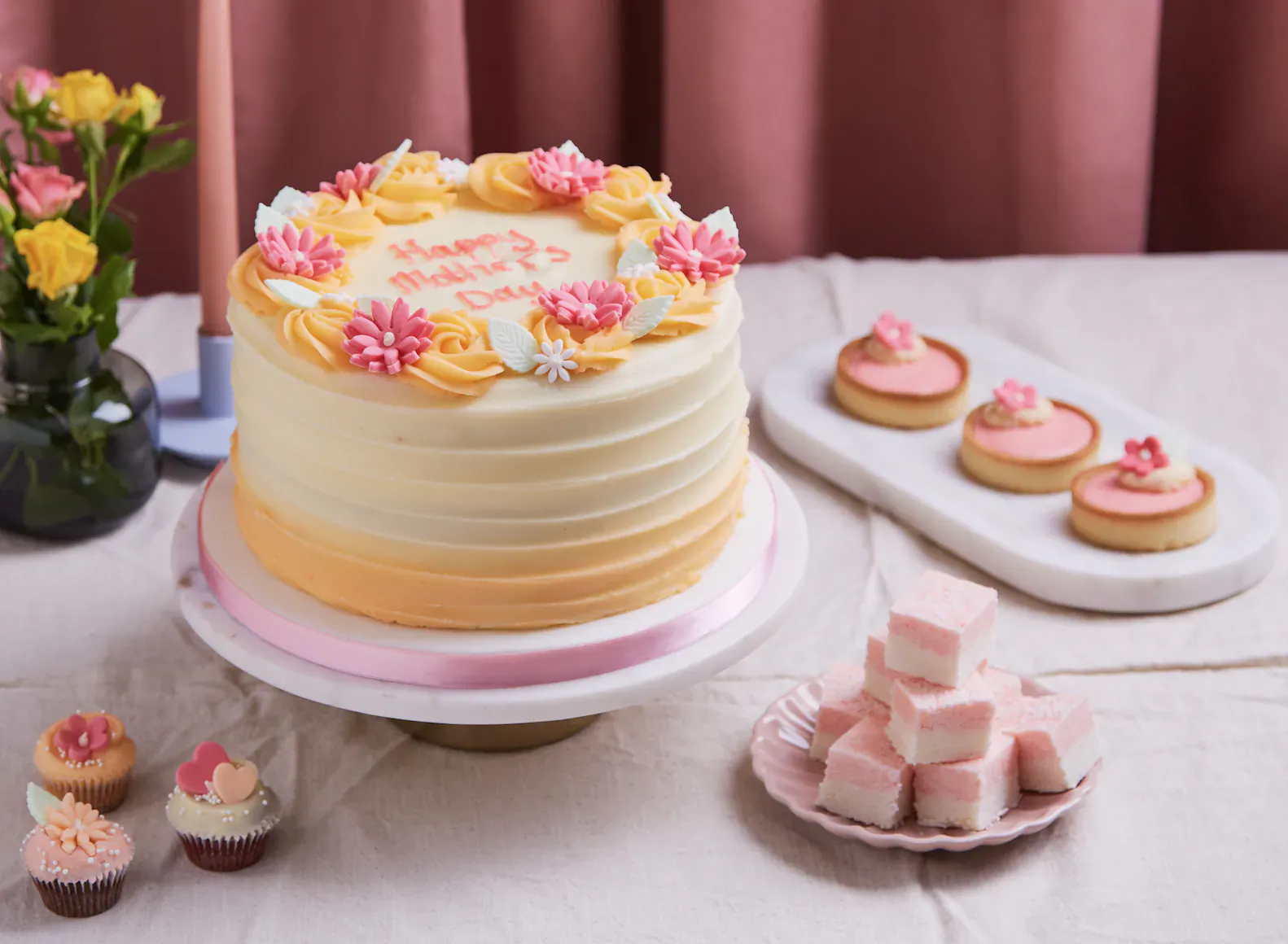 Hummingbird Bakery Mother's Day cake 'Happy Mother's Day' in yellow and pink flowers on white table with macarons and marshmallows