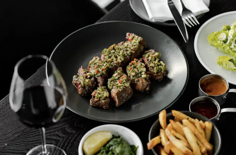 Grilled steaks topped with green herb sauce on black plate, with fries, salad, lemon, sauces, and red wine glass on table.