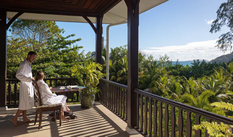 Couple in white robes on a wooden balcony at La Cigale Estate, overlooking lush tropical greenery and ocean.
